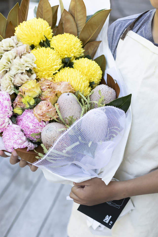 Florist in a pale apron holding a wrapped bouquet of yellow chrysanthemums, carnations and banksia pods, tagged and ready to go out for delivery
