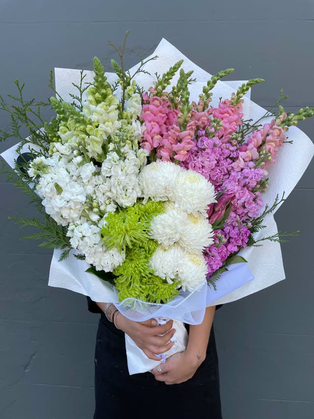 Mixed bouquet of white, pink and purple flowers wrapped in white paper, held in two hands