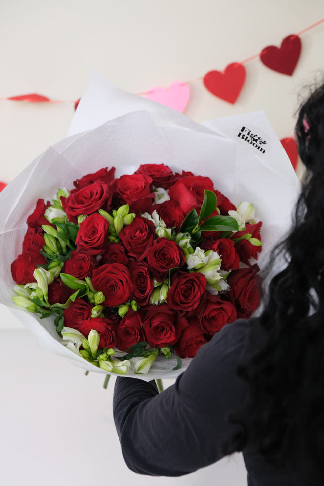A person seen from behind holding a large Fig & Bloom bouquet of red roses and white alstroemeria wrapped in white paper.