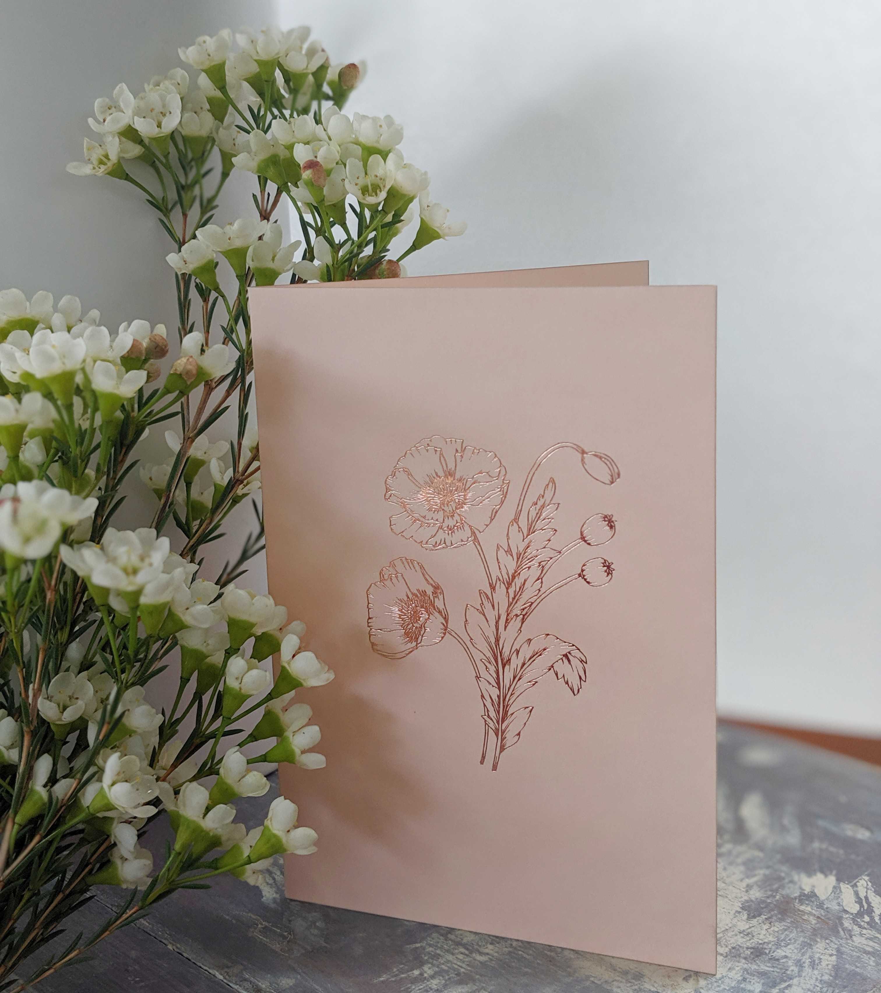 Pink greeting card with a rose gold floral design beside a bunch of white wax flowers