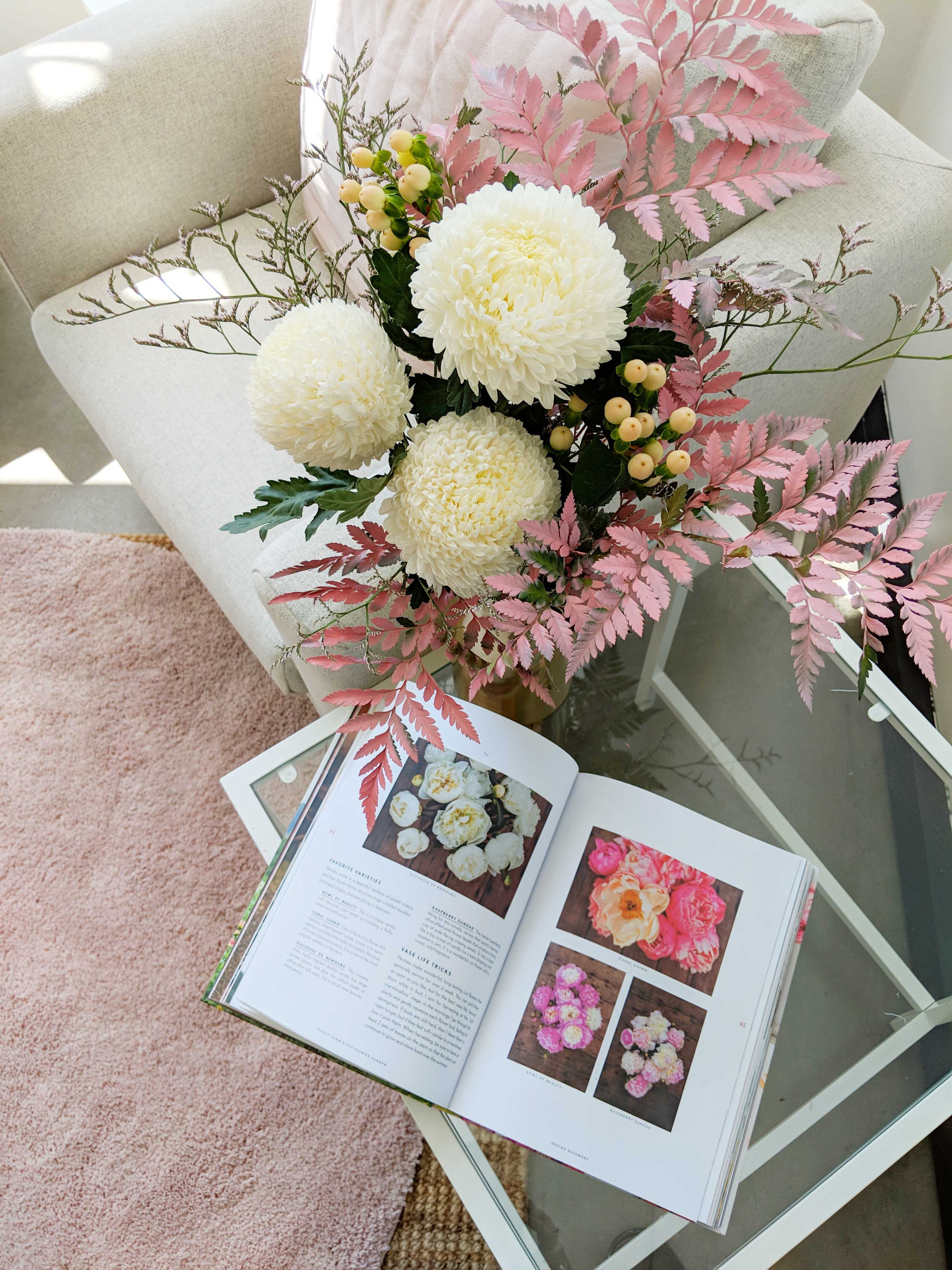 White chrysanthemums and soft pink foliage beside an open book on a coffee table.